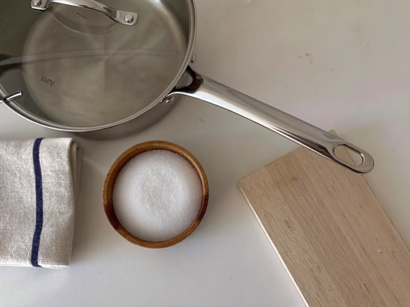 A kitchen counter from above with a stainless pan and glass lid, a folded linen tea towel, a wooden bowl of sea salt and a wooden board